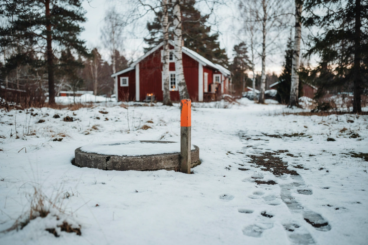 Illustration: brunnslock till enskilt avlopp markerat med käpp i snön vid ett fritidshus i Roslagen